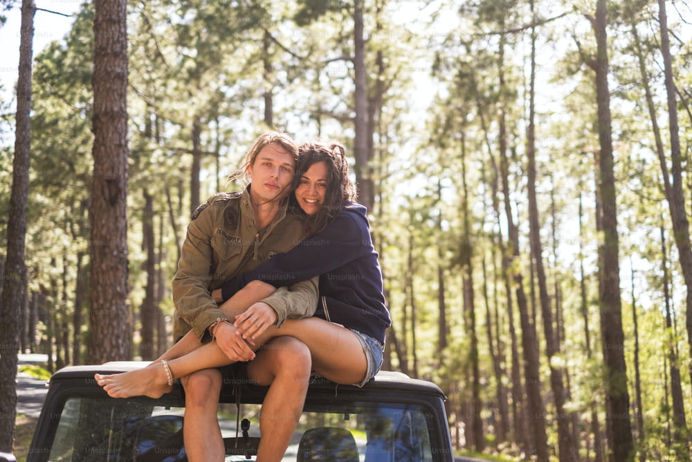 couple in friendship and love sitting on the roof of the car during a travel vacation. parked in the forest with high pines and trees on background. enjoying nature and outdoor leisure activity.