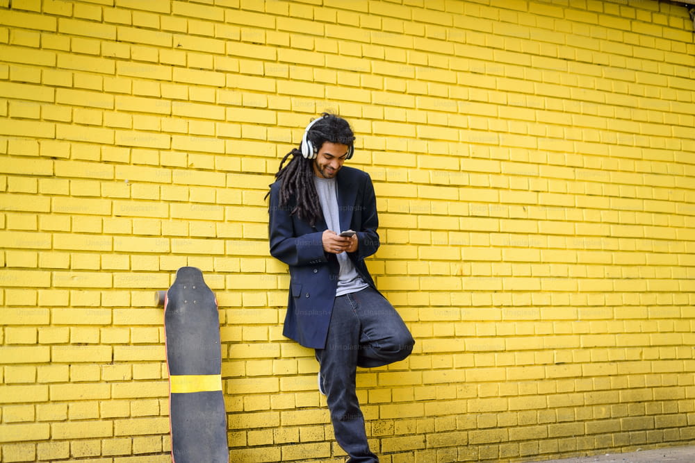 Young cute hipster dreadlocks man leaned on yellow wall listening music with headphones on his head with his long board near him.