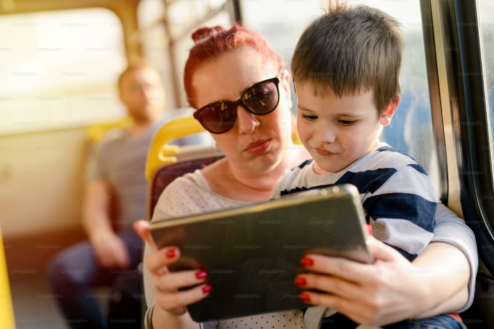 버스에 아들과 함께 앉아 있는 젊은 돌보는 어머니. 소년은 그녀의 무릎에 앉아 태블릿을보고 있습니다.