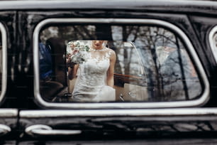 stylish wedding couple sitting in beautiful black car, creative view from window. elegant groom and bride walking and holding hands in city street, wedding day concept