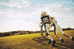 Deux joueurs de football américain s’entraînent à des tacles avec un traîneau de plaquage à l’extérieur sur un terrain de sport en fin d’après-midi