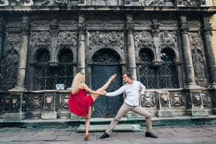 日当たりの良い通りの古い建物で抱き合って踊る愛の美しい幸せなカップル。スタイリッシュなヒップスターの花婿と赤いドレスを着たブロンドの花嫁がダンスを披露し、プロのダンサーが踊ります。ロマンチックなひととき