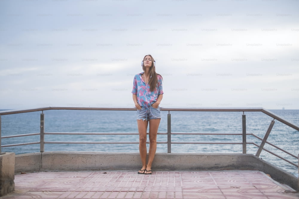 Ganzkörperporträt einer schönen jungen Frau, die mit dem Meer und Wasser blau im Hintergrund steht und den Himmel mit Kopfhörern Musik hört. Modernes Konzept und Freizeitangebot für nettes Mädchen am Strand