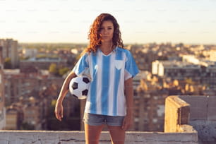 Giovane donna che indossa una maglia da calcio in piedi sul tetto di un edificio, tenendo in mano una palla e guardando un tramonto sulla città.