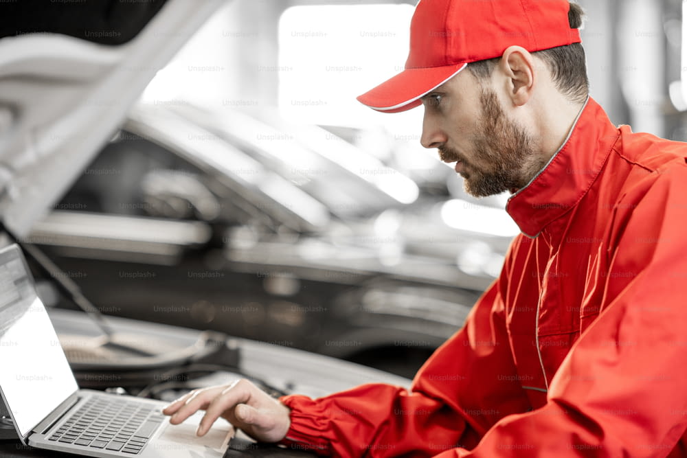Handsome auto mechanic in red uniform doing engine diagnostics with computer in the car service