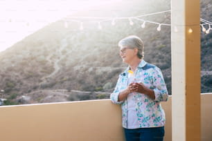 White hair aged beautiful woman drink a cup of white wine outdoor in the terrace looking down and enjoying the sunset at home on the rooftop with mountain view