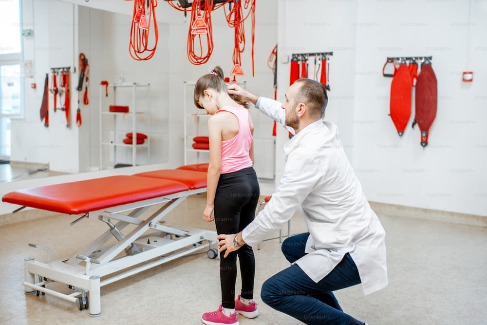 Physiotherapist looking on the back of the junior girl during the medical examination at the rehabilitation office with suspension medical equipment