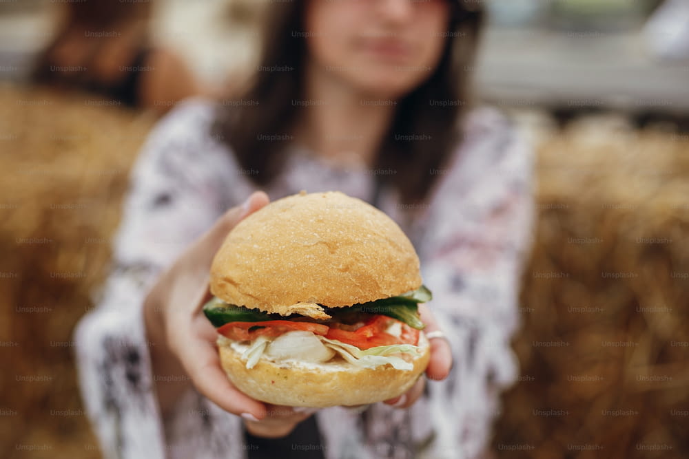 보헤미안 소녀 손에 비건 버거 근접 촬영. 길거리 음식 축제에서 맛있는 비건 버거를 먹는 선글라스를 쓴 세련된 힙스터 소녀. 행복한 보헤미안 여자 시음과 무는 햄버거