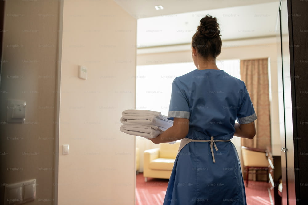 Back view of young brunette chamber maid in uniform carrying stack of clean white towels for guests while entering one of rooms