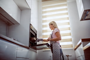 Vista angolare bassa dell'attraente casalinga bionda caucasica in grembiule che tira fuori la cena al forno dal forno.