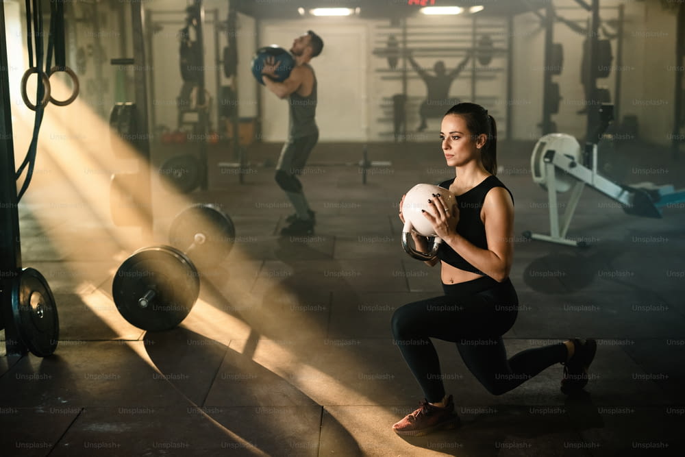 Athletic woman doing split squats while exercising with kettle bell in a gym.