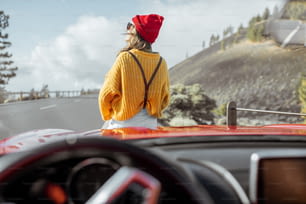 Retrato de estilo de vida de una mujer despreocupada vestida casualmente con suéter brillante y sombrero sentada en el capó del automóvil, disfrutando de un viaje por carretera en la carretera de montaña, vista a través del parabrisas