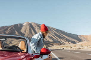 砂漠の渓谷をカブリオレで旅する赤い帽子をかぶった女性が、背景の絵のように美しい火山の近くの道端で車を降りる
