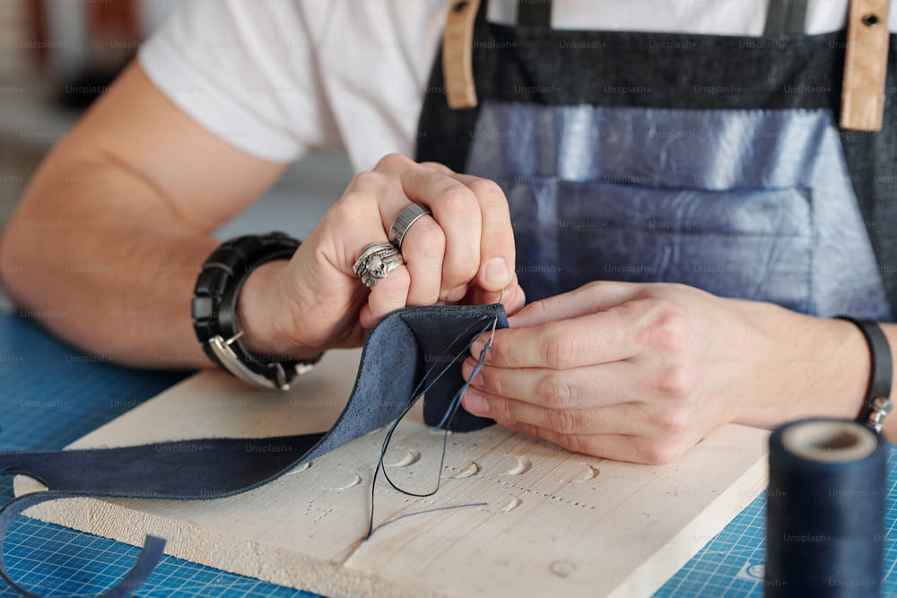 Creative handicraft master with needle holding small piece of black suede over wooden board on table while sewing something