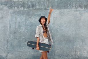 Skater Girl With Skateboard Portrait. Asian Teenager In Casual Outfit Posing Against Concrete Wall At Skatepark. Urban Subculture And Skateboarding As Lifestyle Of Active Teens In City.