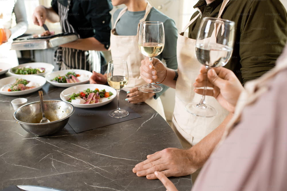Gruppe von Menschen in Schürzen toastet mit Flöten von Champagner oder Weißwein über einem großen Küchentisch mit appetitlichen hausgemachten Speisen