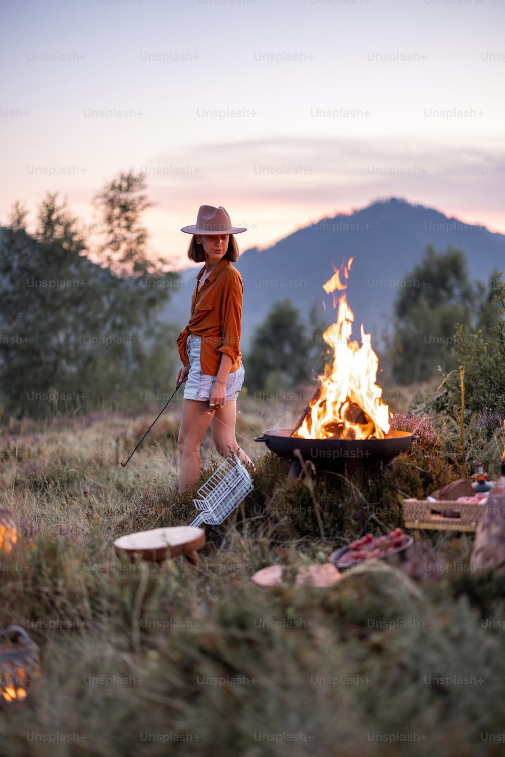 Stylish woman having a beautiful picnic with bonfire and great view on the mountains on the evening. Enjoyment of nature and traveling in the mountains concept