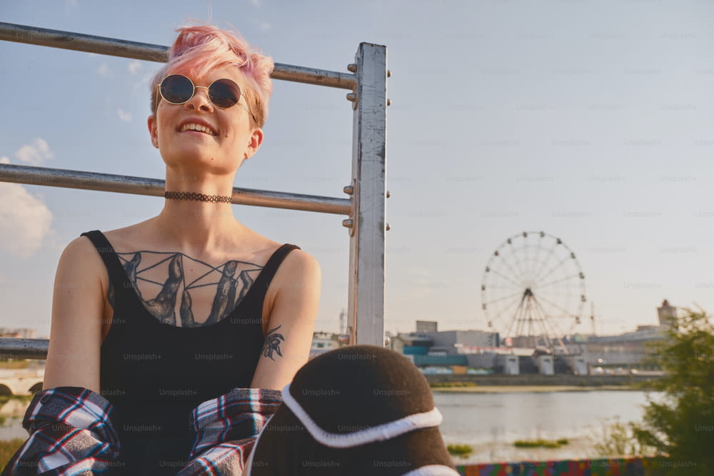 Portrait of daring young tattooed woman with short pink hair, open chest, sitting outdoor in round glasses