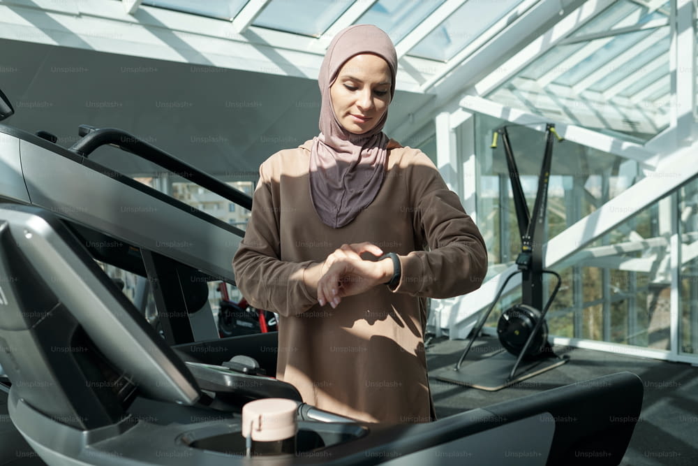 Horizontal medium portrait of modern young adult Muslim woman wearing hijab standing on treadmill in gym checking something on her activity tracker
