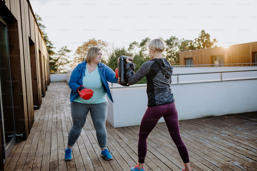 An overweight woman training boxing with personal trainer outdoors on terrace.