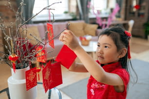 Pretty little girl hanging handmade postcards with new year wishes on branches before holiday