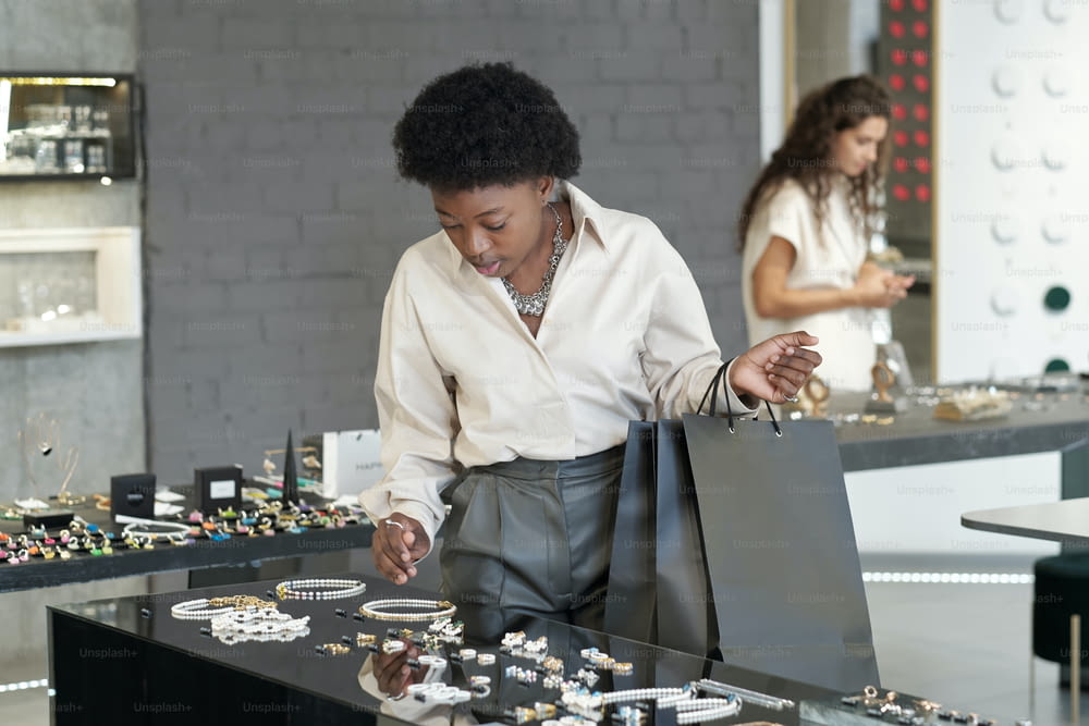 Young African female shopper with paperbags choosing new necklace on large display while looking at jewelry assortment