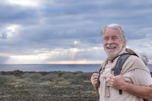 Felice nonno anziano che si gode un'escursione all'aperto in mare con lo zaino in mano. Orizzonte sopra l'acqua e cielo drammatico con il raggio di sole