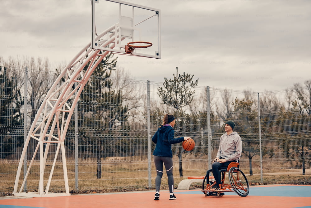 Deportista feliz con discapacidad usa silla de ruedas mientras juega baloncesto con una amiga al aire libre. Espacio de copia.
