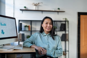 スマートカジュアルウェアを着たアジアの若い女性エコノミストが職場に立ち、コンピューターモニターと財務データを画面に表示