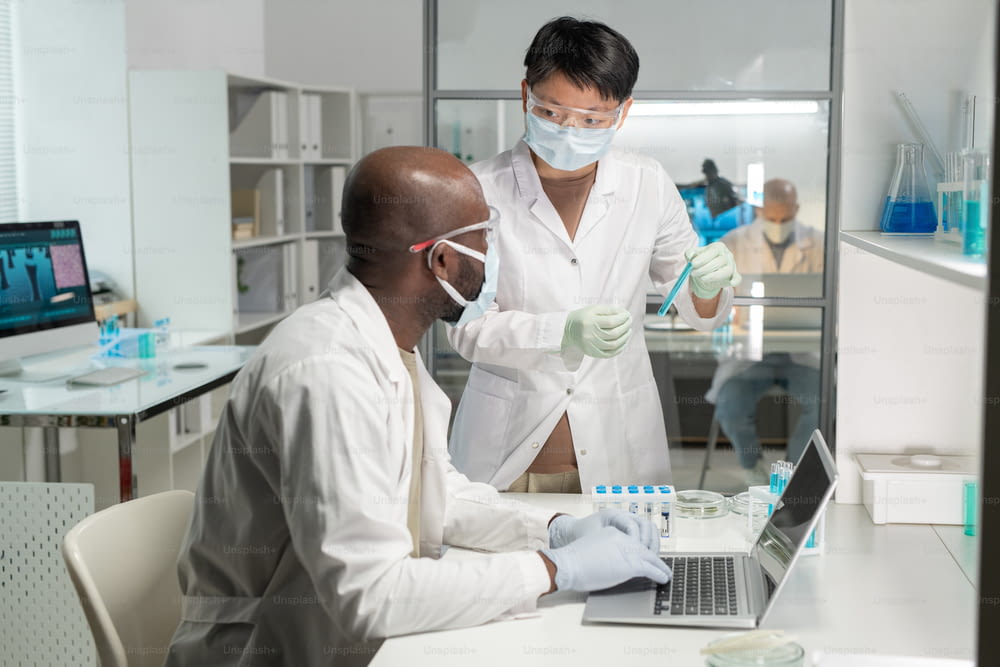 Junge asiatische Wissenschaftlerin in Schutzmaske und Laborkittel zeigt ihre afroamerikanische männliche Kollegenflasche mit blauer Flüssigkeit