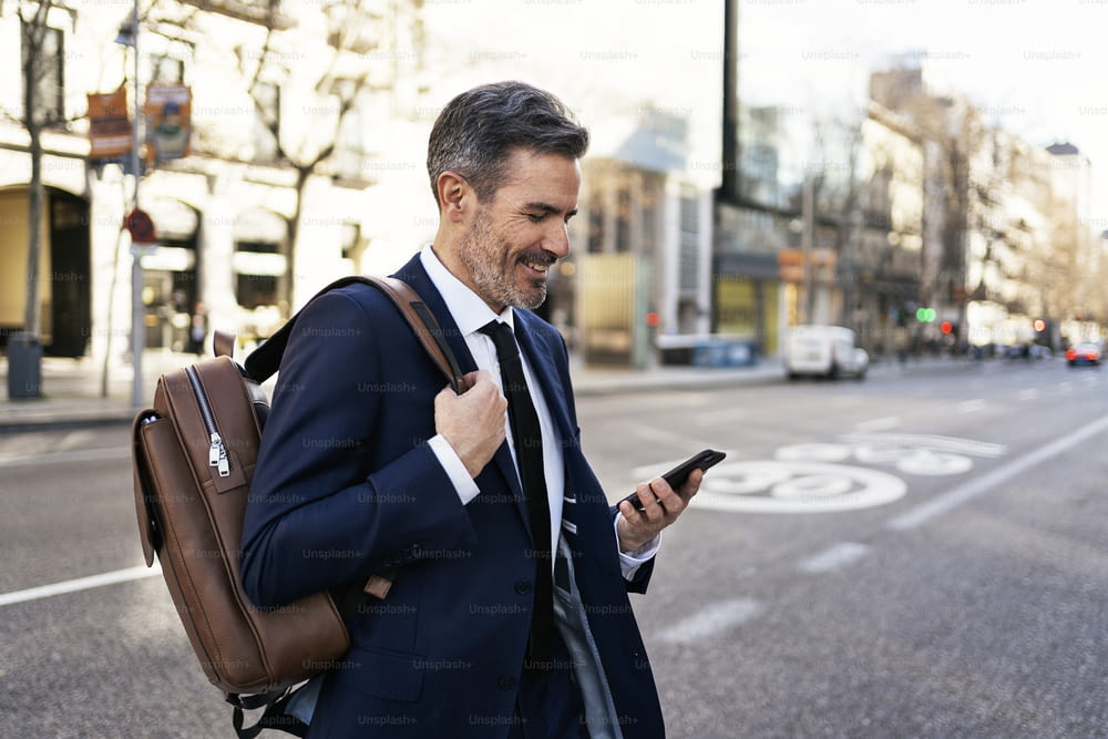 Side view of content mature male entrepreneur in suit and with backpack crossing road and reading message on mobile phone