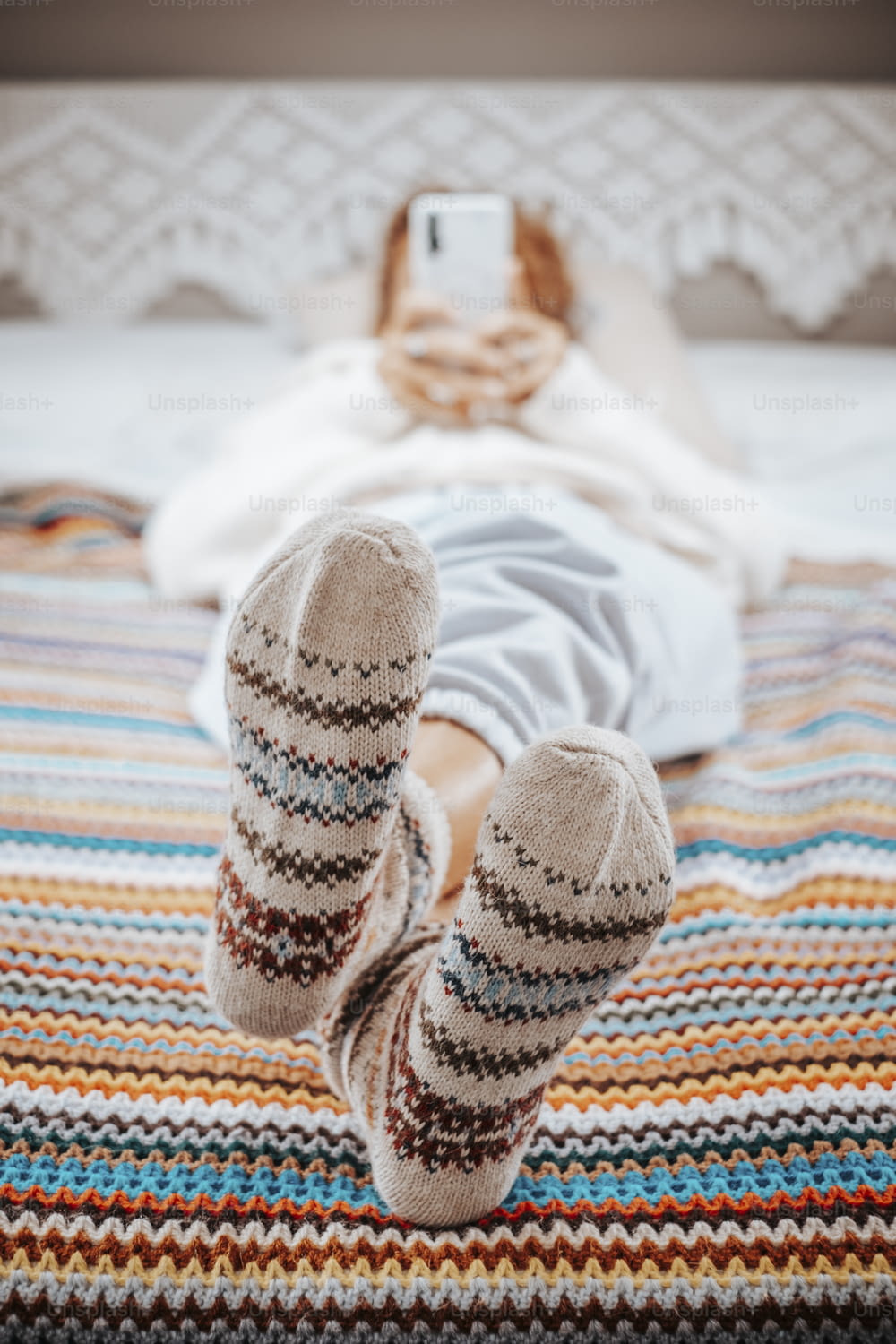 Primer plano de pies y bonitos calcetines en una manta colorida de lana de ganchillo de la misma textura. Mujer que tiene una actividad de ocio relajante en el dormitorio y usa el teléfono móvil en un fondo desenfocado.