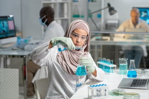 Young serious Muslim female researcher in hijab, gloves and protective eyewear pouring liquid substance into flask with blue fluid