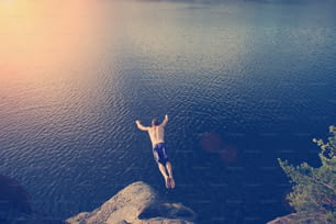 Junger Mann springt von der Klippe in den Fluss (Mann in Bewegungsunschärfe, absichtliche Sonnenblendung und Vintage-Farbe)