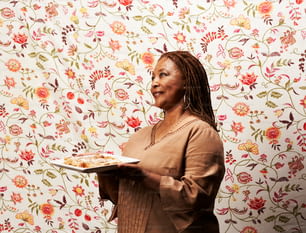 a woman holding a plate of food in front of a floral wallpaper