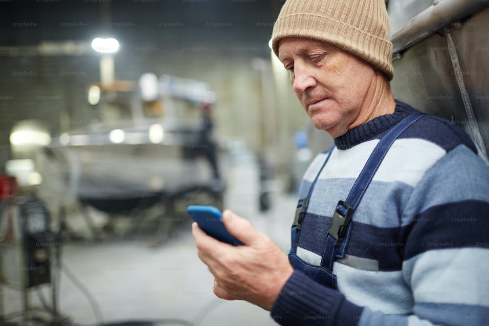 Mature engineer in workwear messaging or reading notification in smartphone