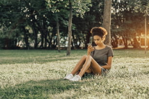 Young charming Brazilian female with curly Afro hair is sitting on the grass lawn in the public park, leaning against the tree and posting her selfies into social networks using the smartphone