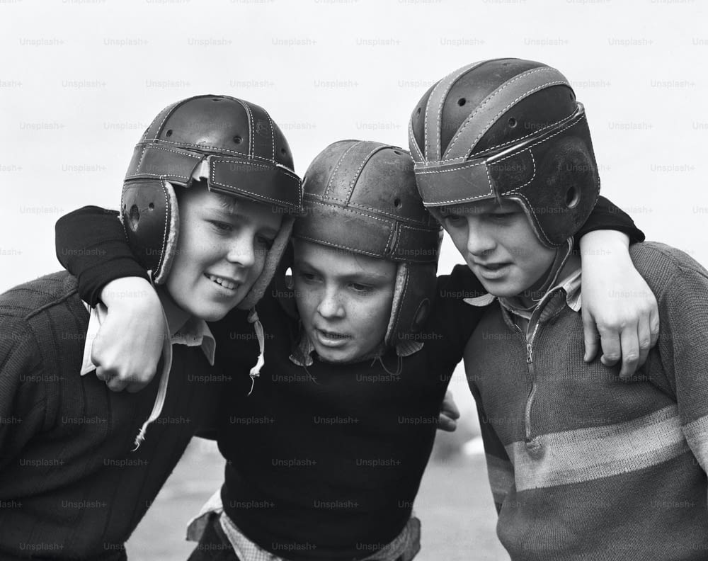 VEREINIGTE STAATEN - CIRCA 1950er Jahre: Drei Jungen mit American-Football-Helmen kauern zusammen.