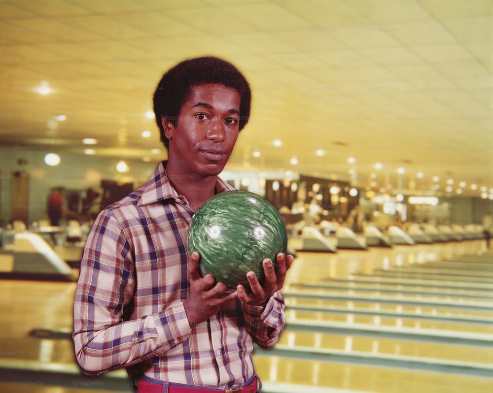 ESTADOS UNIDOS - CIRCA 1970s: Hombre sosteniendo una bola de boliche de mármol verde en una bolera, carriles en el fondo.