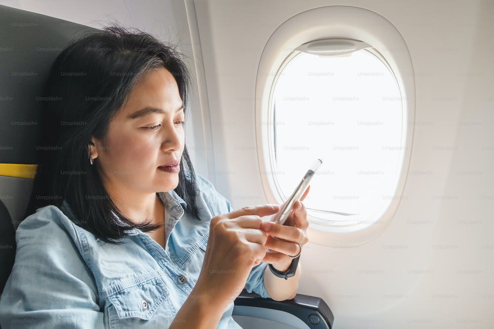 Asian Woman sitting at window seat in airplane and turn on airplane mode on mobile phone before take off.