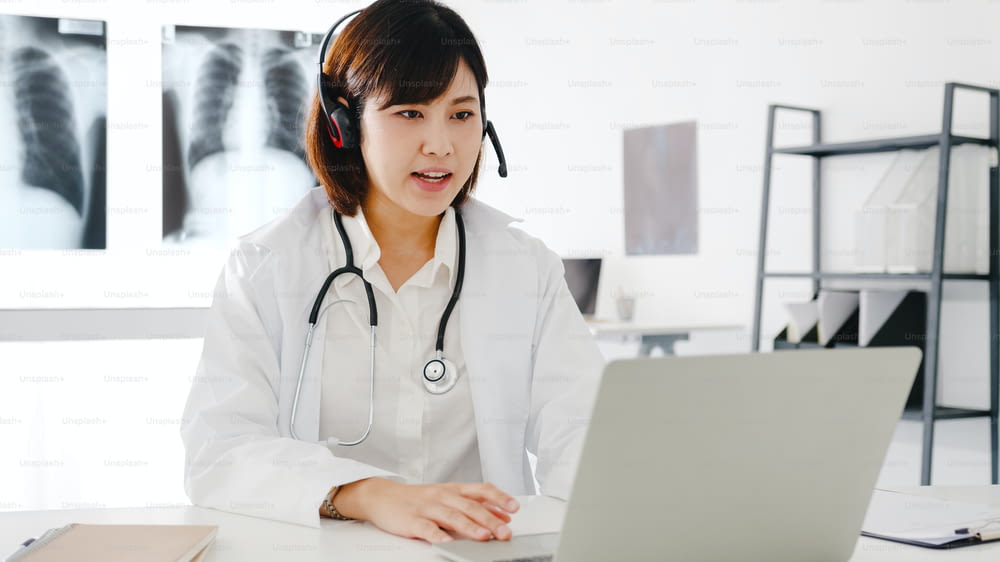 Young Asia female doctor in white medical uniform with stethoscope using computer laptop talking video conference call with patient at desk in health clinic or hospital. Consulting and therapy concept