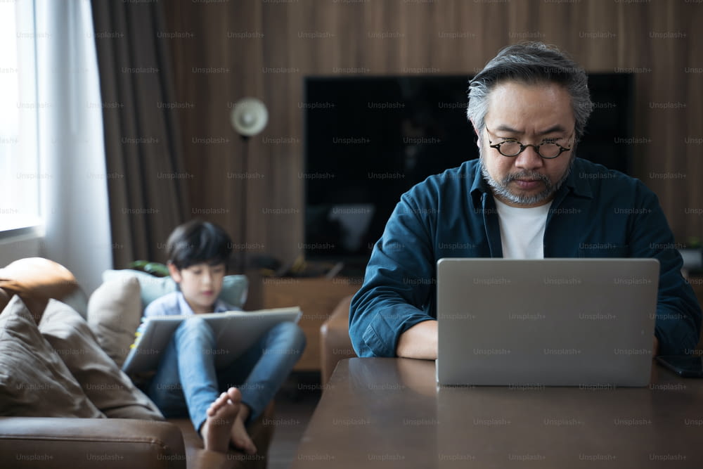 Middle-aged Asian man using laptop work from home with son draws a picture on the back.