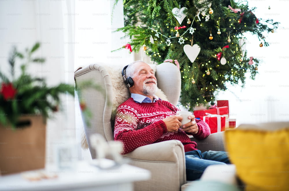 A portrait of senior man sitting with headphones on armchair at home at Christmas time, listening t o music and holding a white cup.