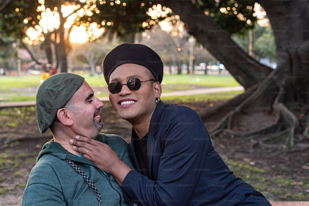Casal gay se abraçando, conversando e rindo enquanto está sentado em uma praia em um parque. Um deles é olhar para a câmera