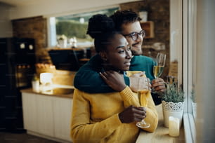 Happy mixed-race couple embracing while having glass of Champagne by the window at home.