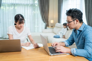 Asian busy Parents working from home due Covid-19 lockdown with children watching TV behind. Business Father and Mother typing laptop and write on copy book didn't pay attention to little daughter.