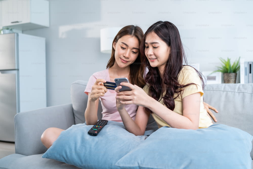 Asian two woman friend enjoy shopping online in living room at home. Beautiful lesbian couple sit on sofa, holding credit card to make e bank online payment after use phone purchase goods in web store