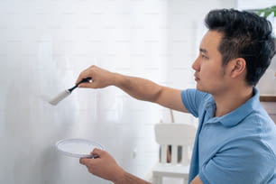 L'uomo attraente asiatico dipinge la parete bianca con il pennello per riparare il soggiorno. Il giovane pittore maschio bello usa il colore del rullo per coprire la parete sporca per rinnovare e decorare la stanza. Concetto di rinnovamento della ristrutturazione della casa