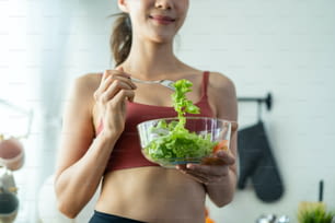 Gros plan d’une femme asiatique attrayante tenant un saladier et mangeant des légumes. Belle fille de sport en vêtements de sport profiter de manger des légumes propres après l’exercice pour la santé à la maison. Concept de régime alimentaire et d’alimentation saine.