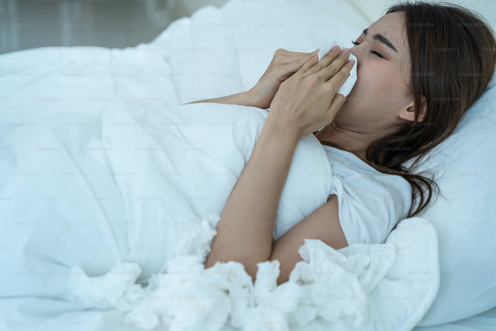 Asian sick girl in pajamas wake up from sleep at night sneezing on bed. Attractive young woman feeling bad and suffer from allergy, put tissue cover her nose while sneezes during bedtime in house.